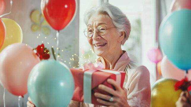 Senior woman with gray hair, wearing an elegant outfit, joyfully opening a birthday gift, surrounded by family, bright sunny room with balloons