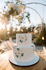 White wedding cake in 2 tiers on a white stand. The cake is decorated with chamomile flowers and green leaves. Cake on the street in the evening at sunset. Glare of the sun. Blurred background