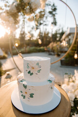 White wedding cake in 2 tiers on a white stand. The cake is decorated with chamomile flowers and green leaves. Cake on the street in the evening at sunset. Glare of the sun. Blurred background