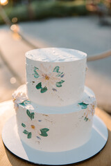 White wedding cake in 2 tiers on a white stand. The cake is decorated with chamomile flowers and green leaves. Cake on the street in the evening at sunset. Glare of the sun. Blurred background