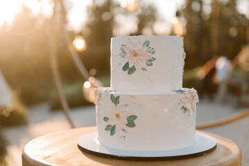 White wedding cake in 2 tiers on a white stand. The cake is decorated with chamomile flowers and green leaves. Cake on the street in the evening at sunset. Glare of the sun. Blurred background