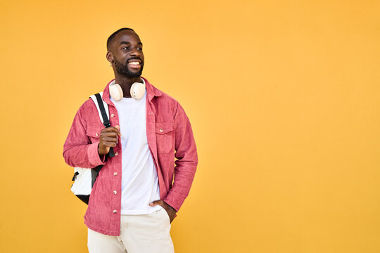 Happy young adult African man hipster standing isolated on yellow background looking away. Smiling generation z Black student guy wearing backpack headphones advertising travel or education offers.