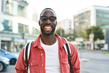 Smiling cool gen z Black hipster guy wearing red shirt, backpack and sunglasses posing outdoors....