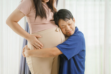 Fototapeta premium An Asian husband sits on a sofa, gently holding his pregnant wife's stomach. He lovingly cares for her as they eagerly await the arrival of their first child, sharing a tender moment.