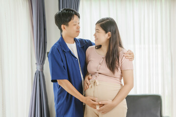 Naklejka premium An Asian husband sits on a sofa, gently holding his pregnant wife's stomach. He lovingly cares for her as they eagerly await the arrival of their first child, sharing a tender moment.