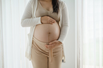 An Asian woman stands while pregnant with her first child, placing her hand on her stomach. She is excited and eagerly waiting for the birth day to meet her baby.