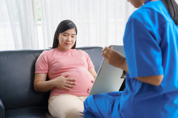 Fototapeta premium Pregnant mothers meet the nurse at the clinic as scheduled, reviewing their medical history and discussing symptoms. Sitting on the sofa, they report headaches, nausea, and back pain to the nurse.