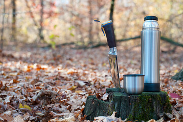 Thermos and hunting knife. The concept of tourism, survival and hunting in the autumn in forest. a thermos of tea, a cup and a knife, on an old tree stump. dry leaves in the park. at nature. close-up