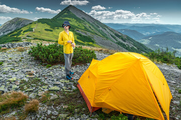 Hiker in yellow shirt standing beside his yellow tent in front of a mountain peak © XArt