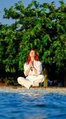 A young and beautiful red-haired girl peacefully meditating by the poolside in a scenic park