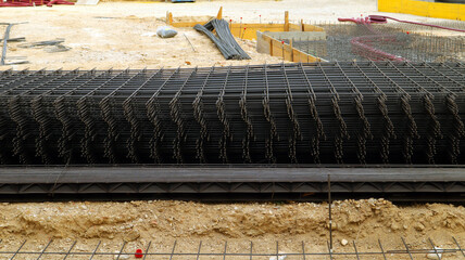 Pile of rebar mesh is stacked on ground at construction site. Mesh is made of black steel and is...