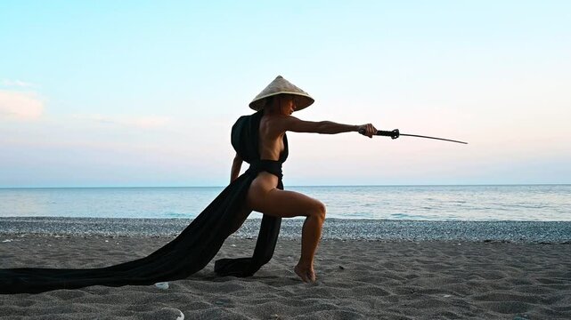 Sexy girl with a gorgeous body, naked in a black kimono and an Asian hat, with a katana in her hand on the seashore in the image of a samurai training with a sword