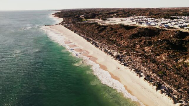 A sandy beach with waves, brown coastline, vegetation, town in distance, and setting sun