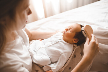 Mother holding infant baby boy sleeping on hands and brushing his hair in room. Motherhood.