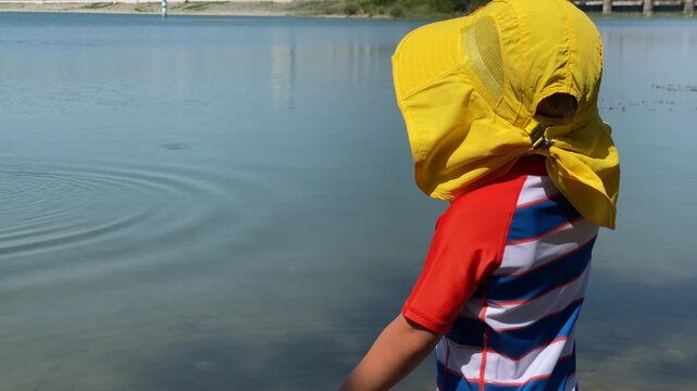 Small child playing on the lake shore on a summer day. Little boy wearing sun protection shirt and funny yellow sun hat in nature. Simferopol, Crimea, Russia 2024.