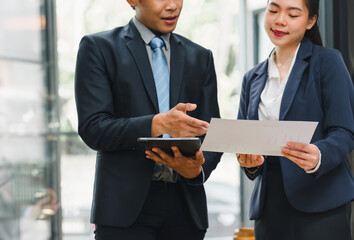 Two business professionals in suits discuss strategy while using a tablet and holding documents, highlighting collaboration and planning.