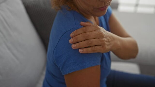 Woman sitting indoors at home holding her painful shoulder in a blue shirt, showcasing discomfort and emphasizing a domestic setting in an interior living room.