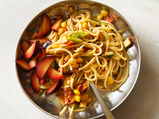 A vibrant mix of vegetable spaghetti, beautifully complemented by juicy plums, served on a shiny steel plate. Fresh herbs and toasted nuts add a final touch of flavor and texture.