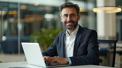 Fototapeta premium A confident businessman with glasses smiles while typing on his laptop in a modern, well-lit office environment..