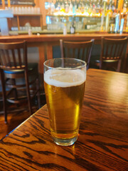 Glass of beer on wooden bar table in dimly lit pub with bar counter and chairs in the background. City life. Happy hour.