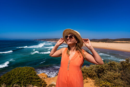 Beautiful woman in fashionable orange dress at viewpoint on cliff near sunny and sandy beach Bordeira, Algarve, Portugal
