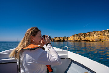 Summer vacation in Lagos Portugal. Woman of European beauty in white sweatshirt and life jacket taking photos of cliffs during boat cruise to Ponta da Piedade on beautiful sunny day. Boat trip to sea