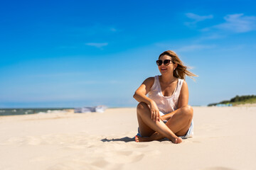 Summer holidays. Portrait of young beautiful woman with long blonde hair in light dress and sunglasses sitting on ground on sandy beach on sunny day. Energy, joy of life and youth