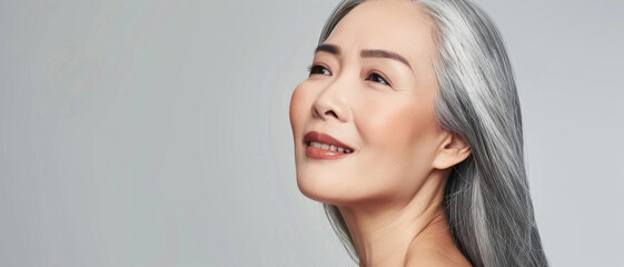 A gracefully aging woman with silver hair gazes upwards with a serene and content expression against a neutral backdrop.