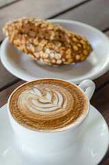 A close-up of a steaming cup of coffee with latte art, served on a wooden table with a pastry on the side.