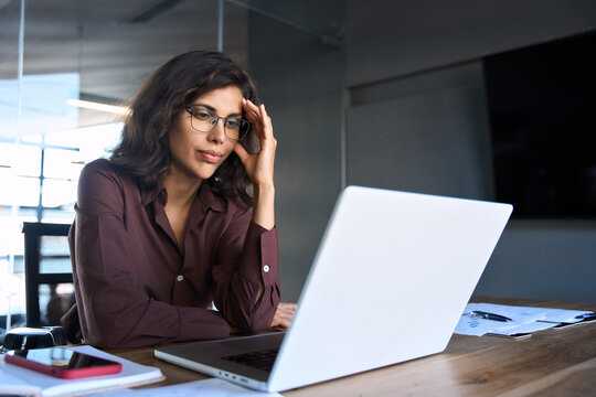 Focused young businesswoman looking at laptop pc computer screen having headache, migraine. Latin business woman holding hand near temples, feeling stressed and tired sitting at workplace in office