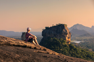 Man with backpack sitting on rock and looking at landscape at sunset. Beautiful scenery with Sigiriya rock. Solo traveler in Sri Lanka. .