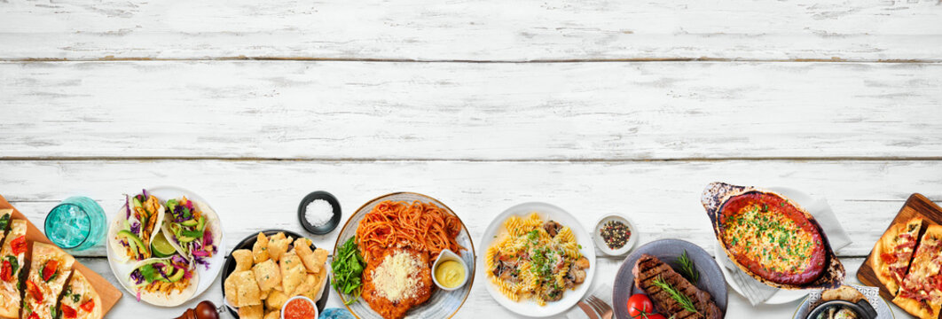 Bottom border of various delicious foods. Top down view on a white wood banner background. Copy space. Pastas, flatbreads, tacos, steak and restaurant theme dishes.