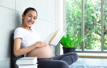 Smiling pregnant woman reading her favorite book, sitting in living room and touching belly on white. Time for hobby and relax. Mother and Baby Free Space.