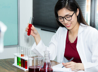 Asian scientist of laboratory researcher holding medical glass tube with red liquid wearing white gown and special lab eyewear. Scientist woman at job. Covid and Chemistry concept