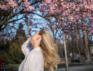 Fashion outdoor photo of beautiful woman with blond hair in elegant suit posing in spring flowering park with blooming cherry tree. Copy space and empty place for advertising text