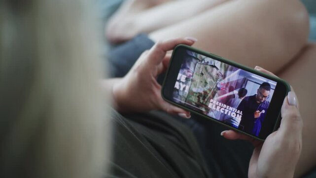 Close up of Caucasian woman holding mobile phone in hands and watching political advertising about the United States presidential elections and voting while lying on sofa at home. Election Day in USA.