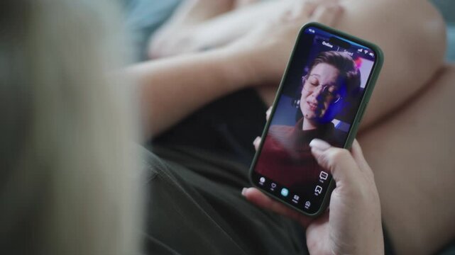 Caucasian woman scrolls through and watches app feed with short videos using mobile phone while lying on sofa. Smartphone application with various video content displayed on phone screen. Close Up.