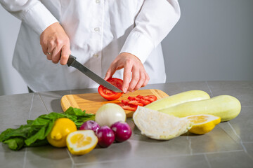 Chef cutting tomatoes on wooden board on grey table, lemons, zucchini, cabbage, onions, green leaves are placed around