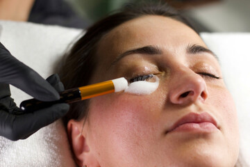 A beautician in black gloves meticulously applies a cleansing product around a woman's eyes using a...
