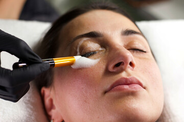 A beautician wearing black gloves uses a brush to apply a cleansing product around a woman's eyes. 