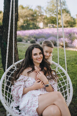 Beautiful young smiling woman mother brunette with son boy child sitting on a wicker hammock, swing in the park and hugging. Photo of a happy family, portrait.