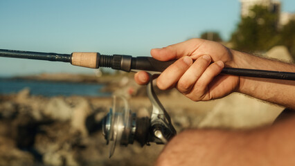 Close-up of a fisherman standing on shore spins the reel on a spinning the fishing rod	
