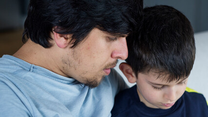 Obraz premium Close-up shot of a father and son sitting on a couch, focused on a tablet computer. Tender moment of family connection and the integration of technology into daily life. 