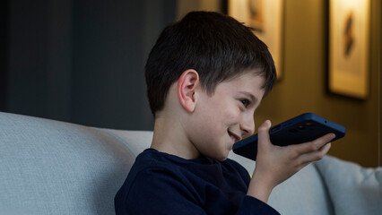 A young boy sits on a sofa, smiling, talks on his smartphone. Modern childhood and digital communication, kid engaged in a cheerful conversation in a cozy home	
