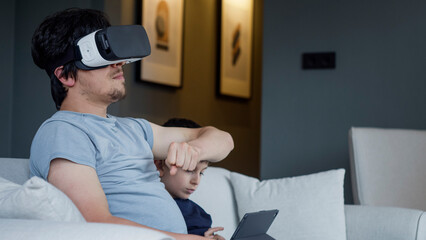 A man immersed in a virtual reality experience with a VR headset. A young boy sits engaged with a tablet. Modern family life, the immersive and interactive of VR technology in a cozy home	
