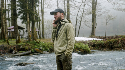 Obraz premium Tourist man talking on the phone near the river. Young hiker in forest camp, using mobile phone, traveling and hiking alone. 