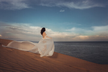 Fototapeta premium Elegant Woman in Flowing White Dress on Coastal Sand Dune: Ethereal Beach Sunset Scene