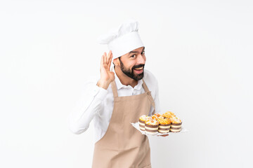 Young man holding muffin cake over isolated white background listening to something by putting hand on the ear
