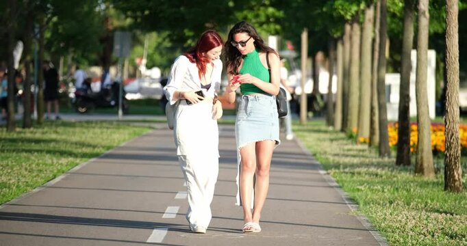 Two young and beautiful female friends are walking together in a park with a tree-lined path, chatting and enjoying their time