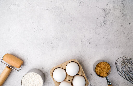 Baking background with cooking ingredients includes flour, sugar, eggs, and a wooden rolling pin on the light stone table.